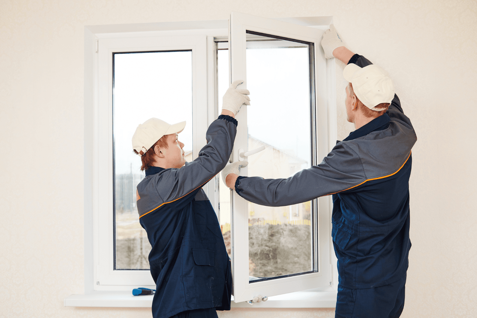 Two workers in uniforms are installing a white-framed window inside a home.
