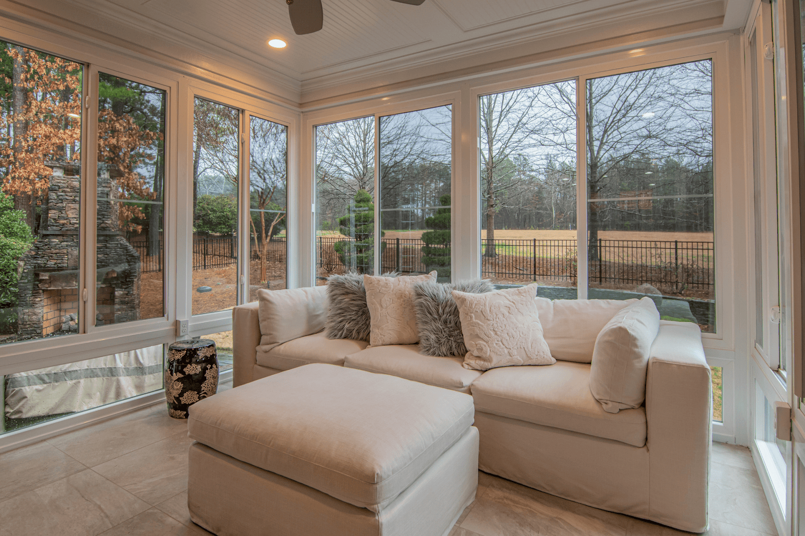 Bright sunroom with large windows, a cream-colored sofa, and outdoor views of trees and a stone fireplace.