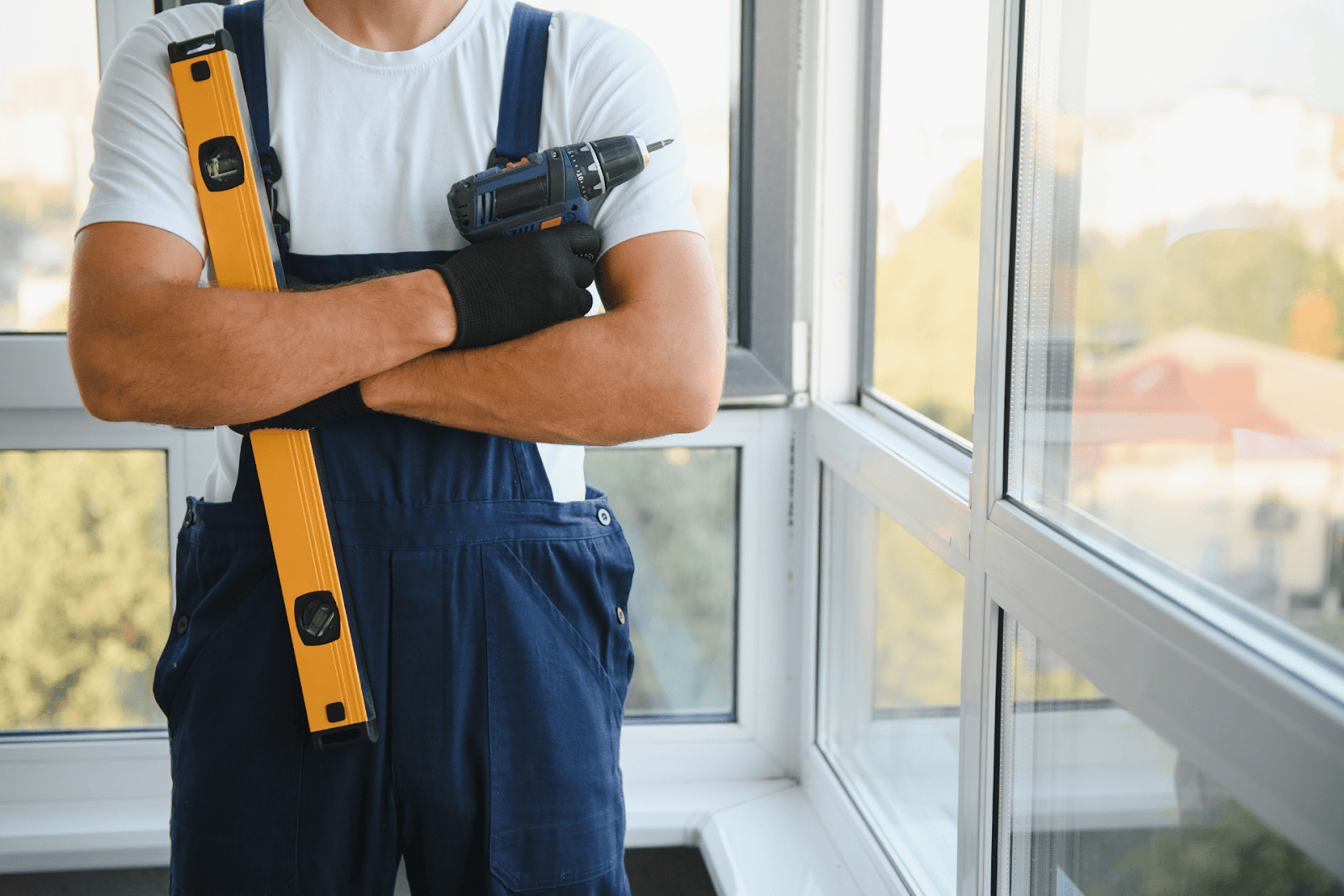 Worker in overalls holding a power drill and level tool near large windows.