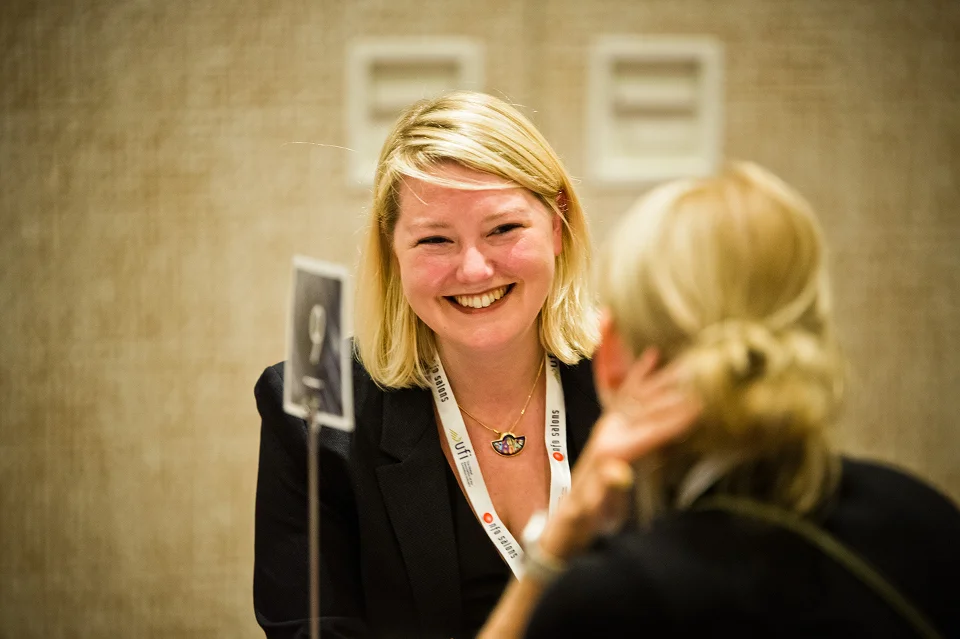 Smiling woman with blonde hair wearing a black blazer and lanyard, engaging in conversation.