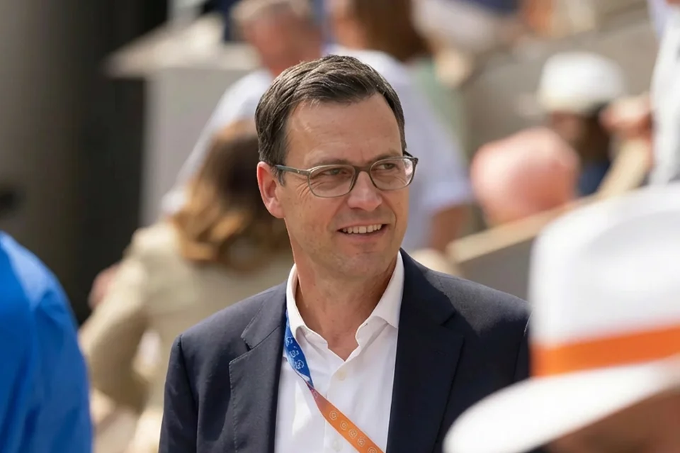 Man with glasses wearing a dark blazer and white shirt in an outdoor setting with blurred background.