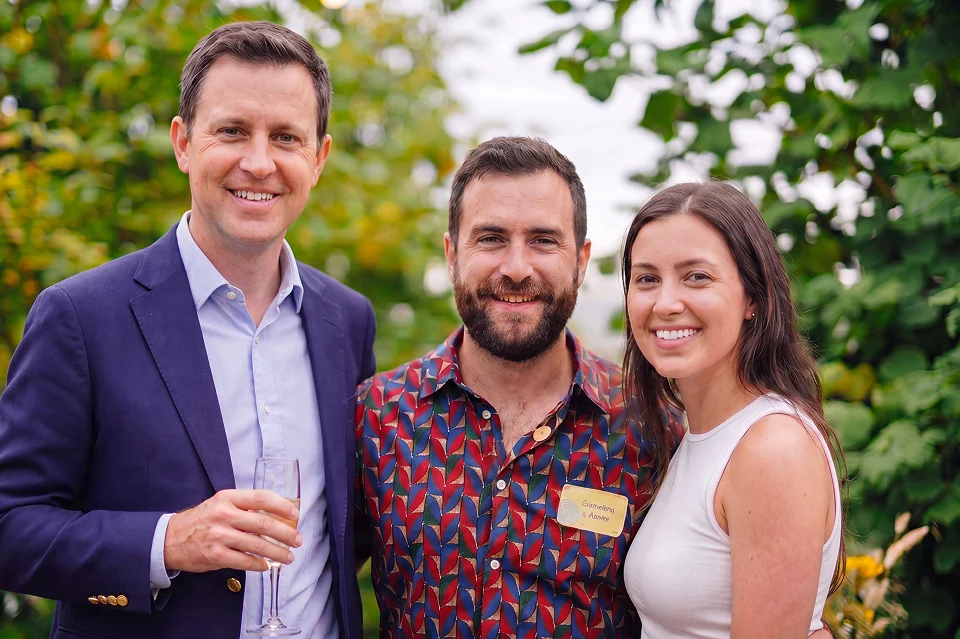 Three people smiling outdoors: a man in a blazer holding a glass, a bearded man, and a woman in a white top.