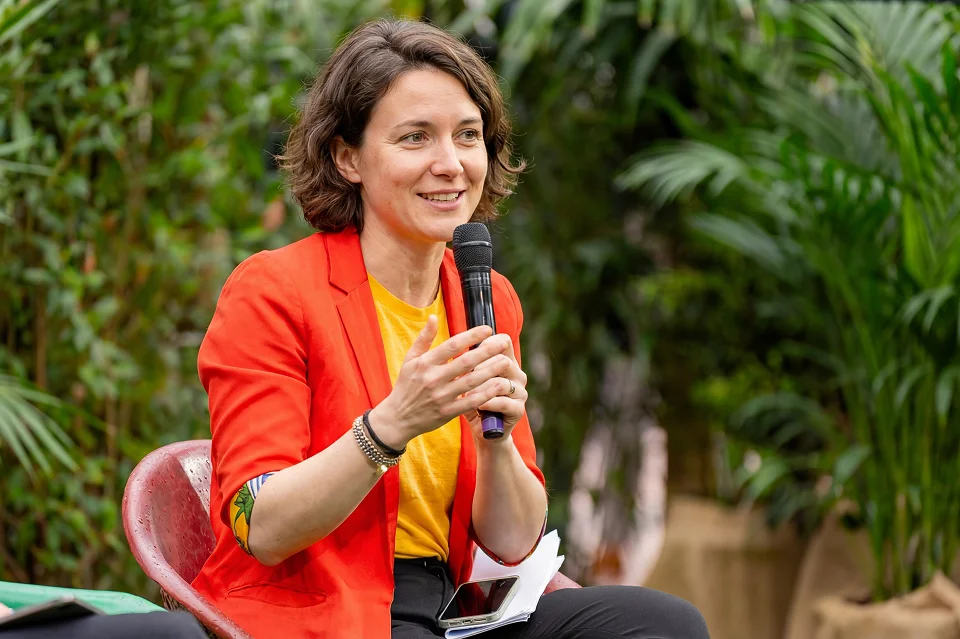 Woman in a red blazer and yellow shirt speaking into a microphone outdoors with plants behind her.