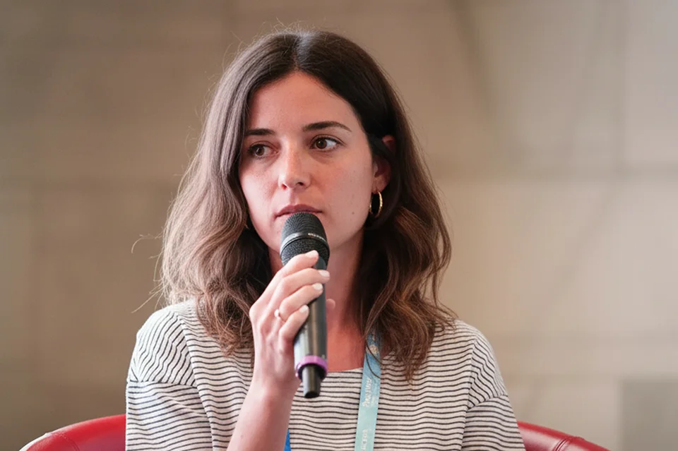 Woman with brown hair holding a microphone, wearing a striped shirt and blue lanyard.