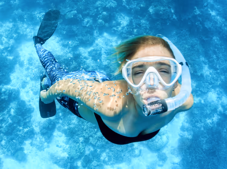 Person snorkeling underwater wearing a mask, snorkel, fins, and patterned swimwear in clear blue water.