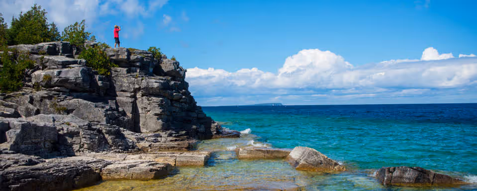 Person in red shirt standing on rocky cliff by clear blue ocean under a partly cloudy sky.