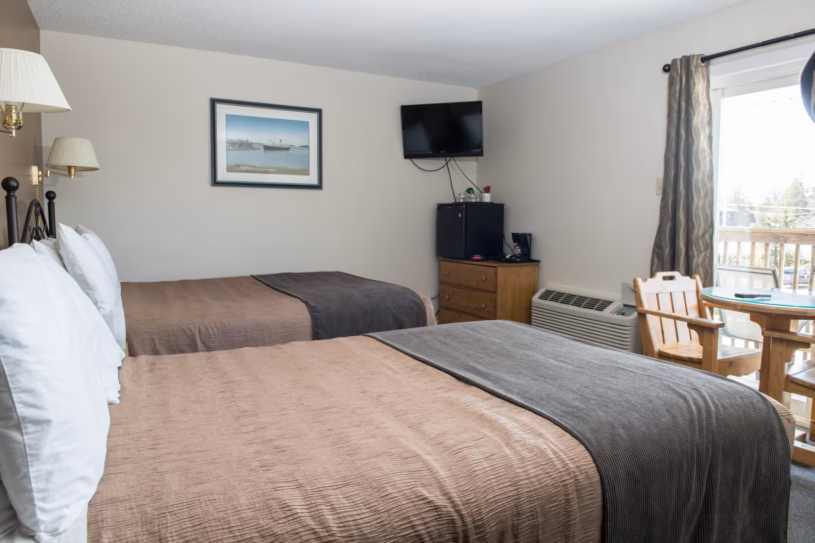 Hotel room with two double beds, a wall-mounted TV, small fridge, and table with wooden chairs near a sliding door.
