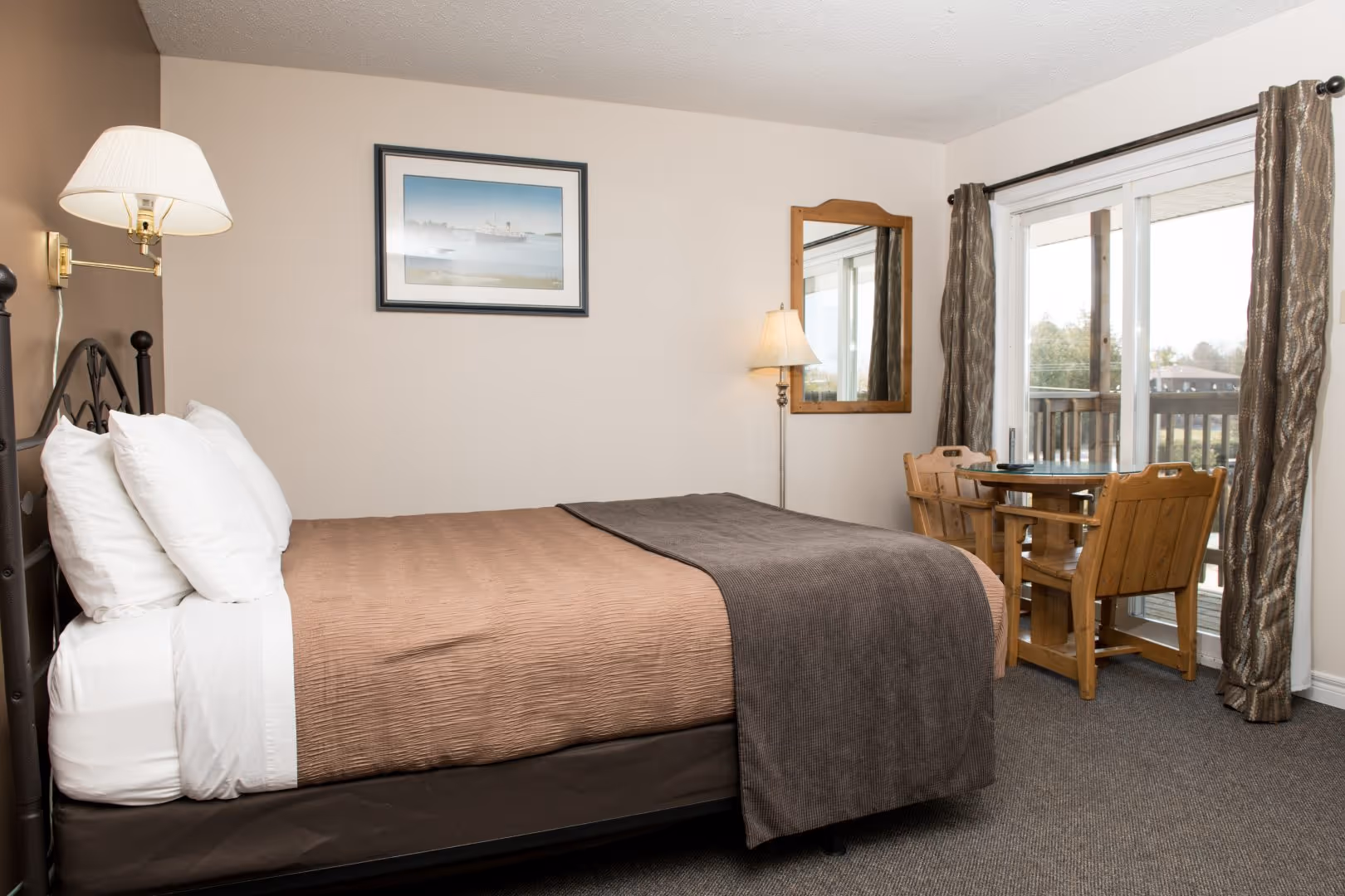 Bedroom with brown bedspread, white pillows, wooden table and chairs, floor lamp, and sliding glass door.