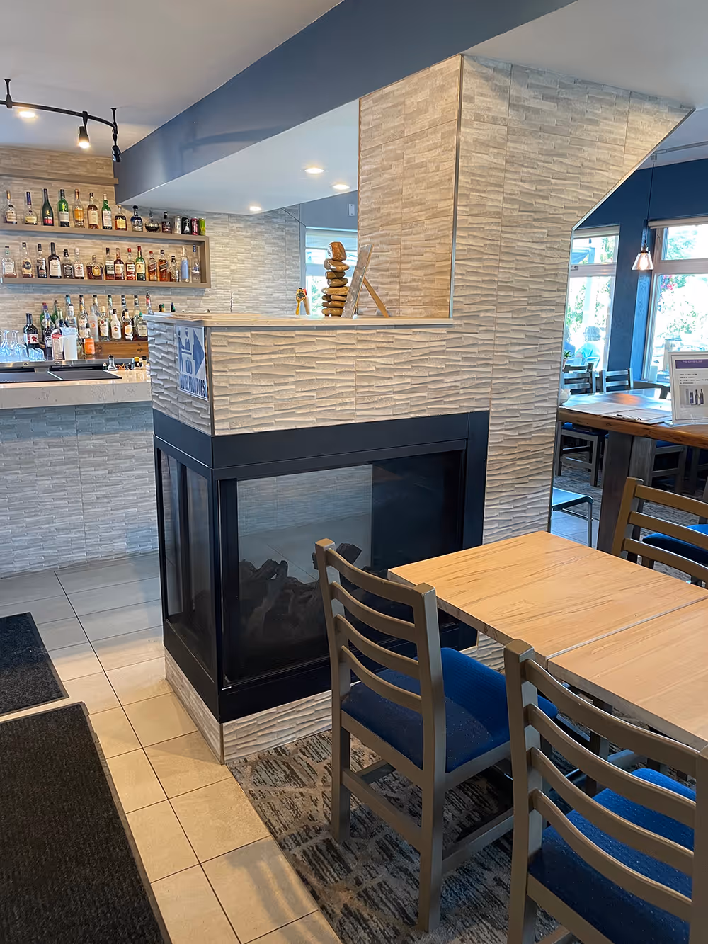 Modern dining area with wooden tables and blue chairs beside a glass-panel fireplace and a stocked bar in background.