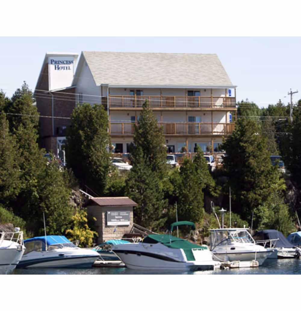 Three-story Princess Hotel behind trees with several boats docked in calm water in front.