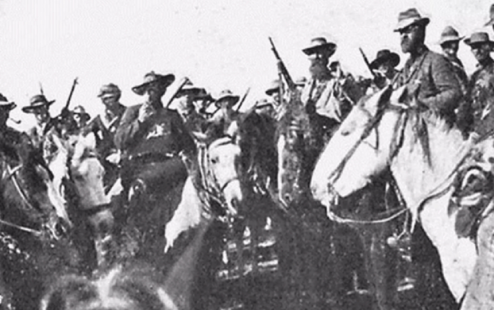 Black and white photo of a group of armed men on horses wearing hats and military-style clothing.