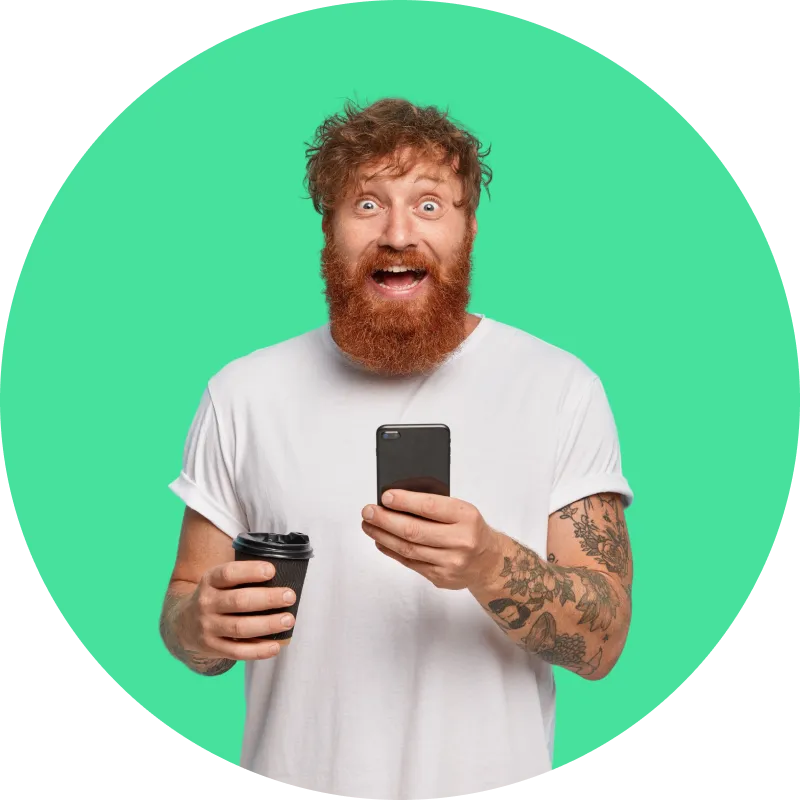 Hombre con barba pelirroja y tatuajes, sonríe mientras sostiene un teléfono y un vaso de café, sobre fondo verde.