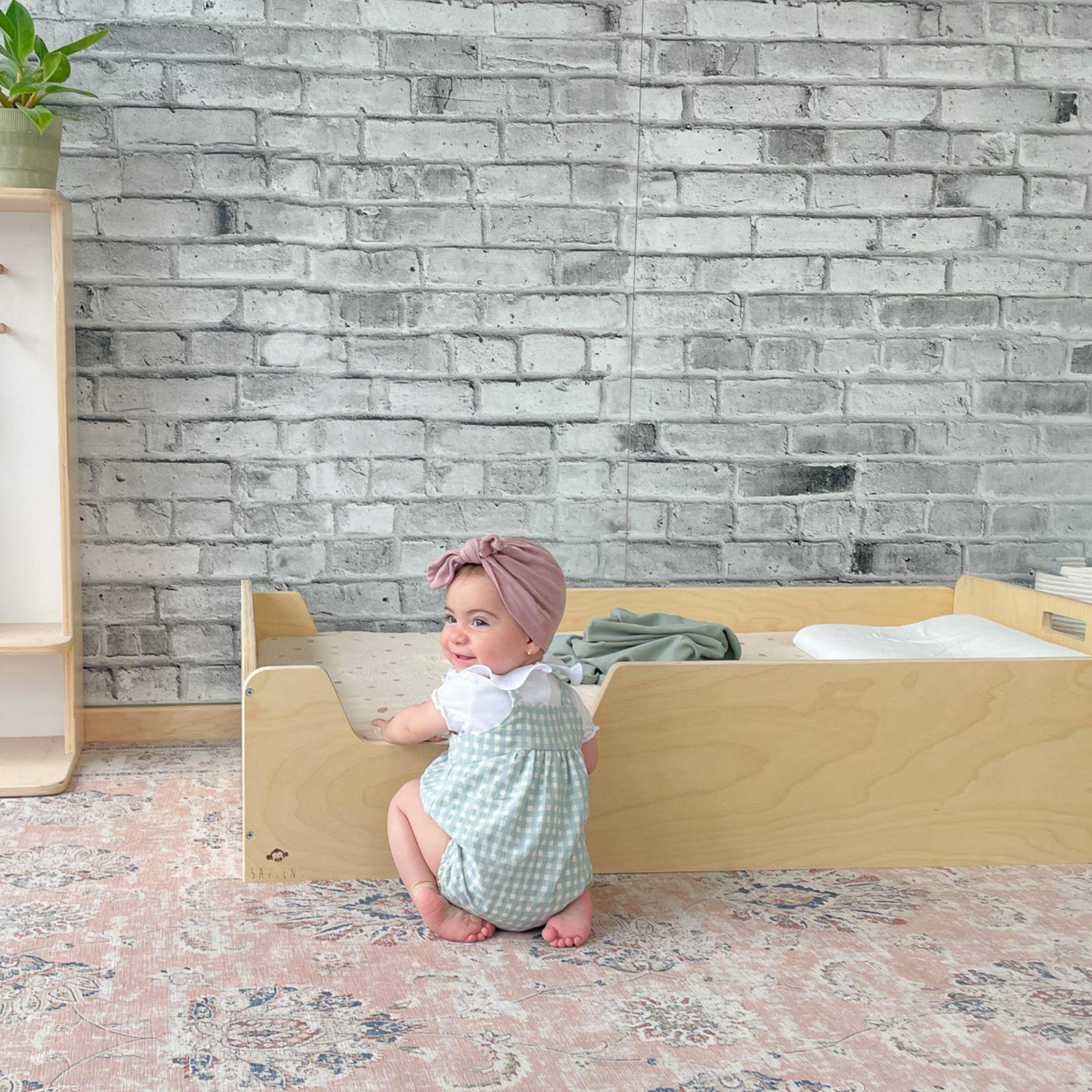 Baby in checkered romper and pink turban sitting by wooden shelf