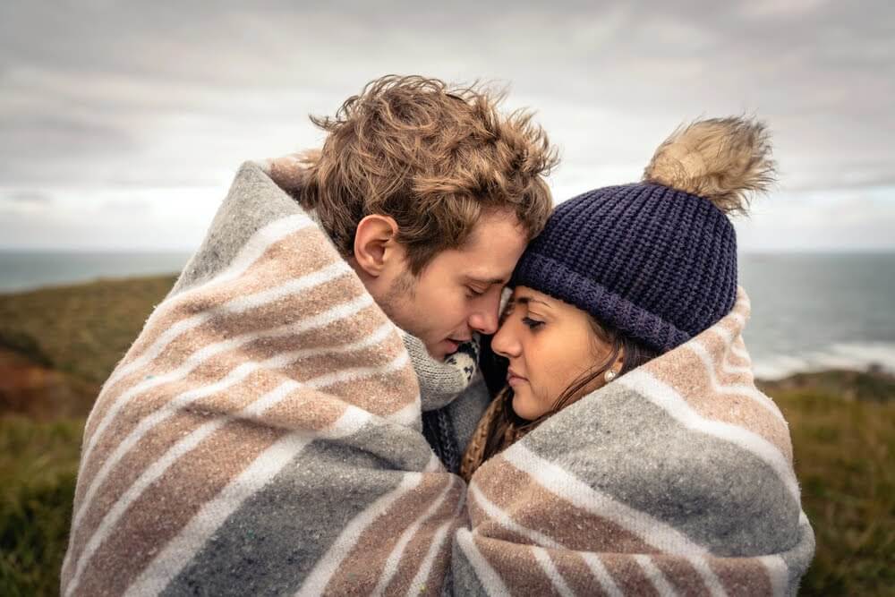 Couple wrapped in blanket, leaning close together by coastal landscape