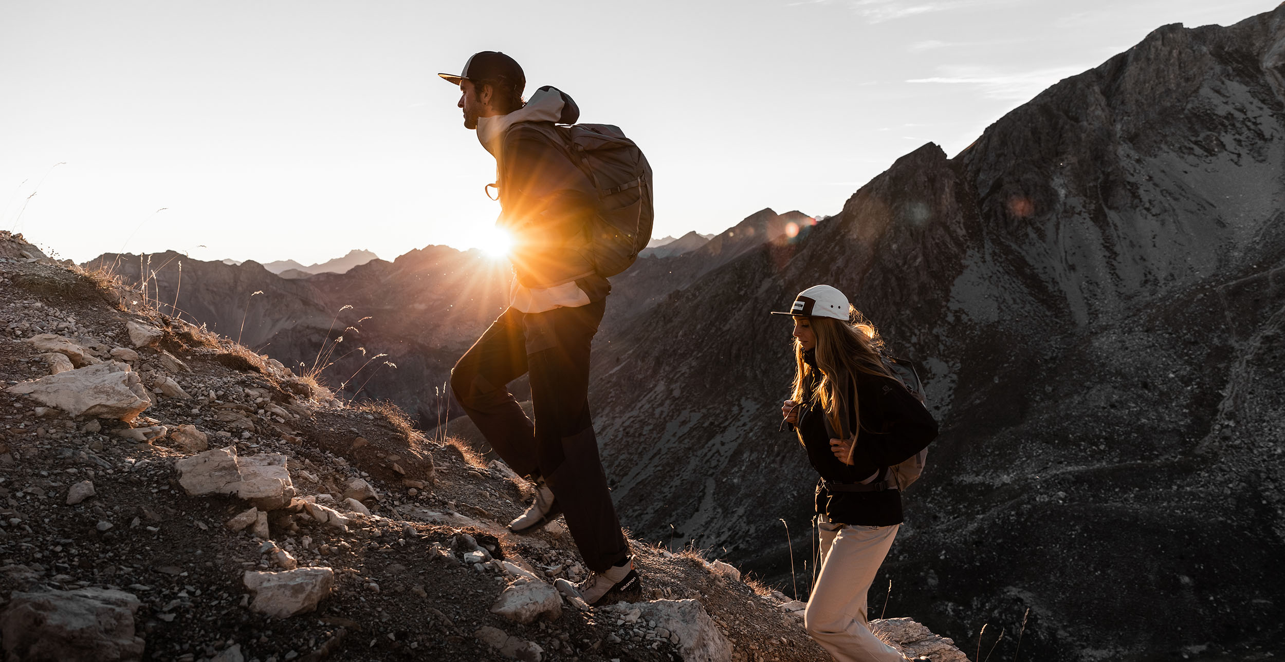 Two hikers climbing rocky mountain trail at sunrise with dramatic backlight