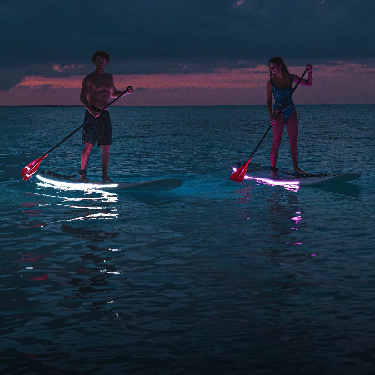 Two paddleboarders glide on illuminated boards at sunset