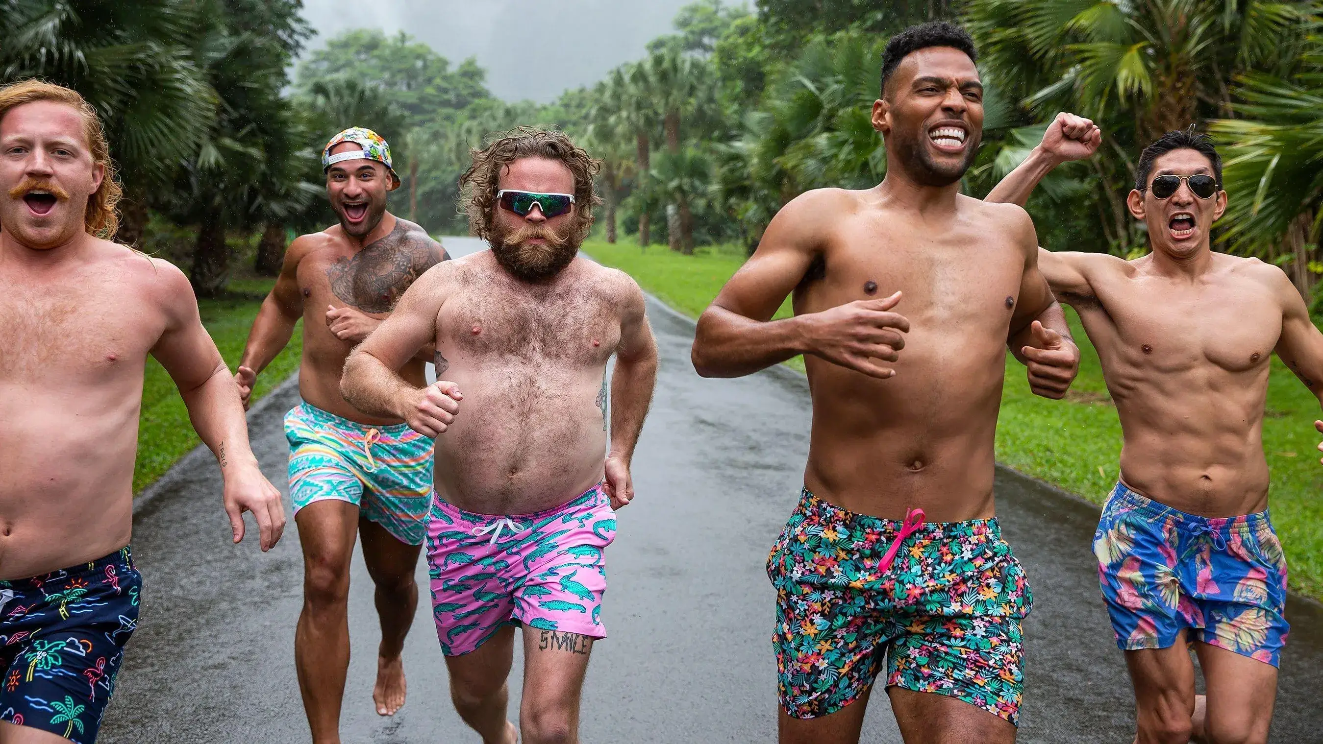 Five shirtless men running together on palm-lined road, wearing colorful shorts