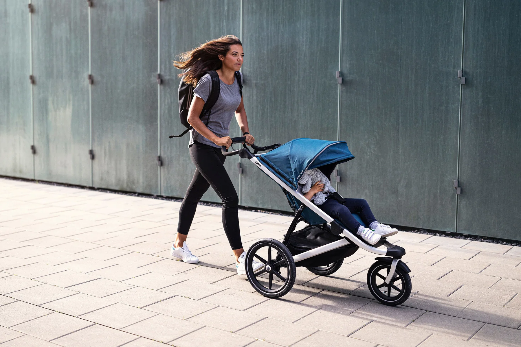 Woman pushing blue jogging stroller while walking on urban pathway