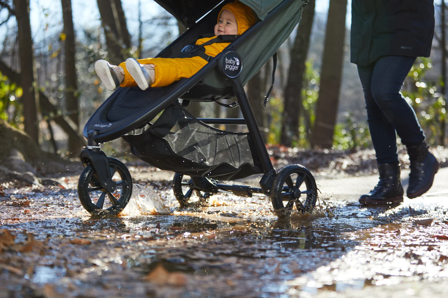Baby in yellow snowsuit enjoying stroller ride through muddy forest puddles
