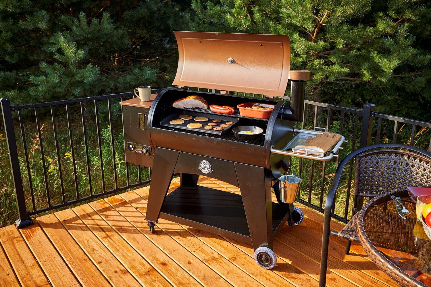 Pellet grill with various foods cooking on wooden deck surrounded by trees