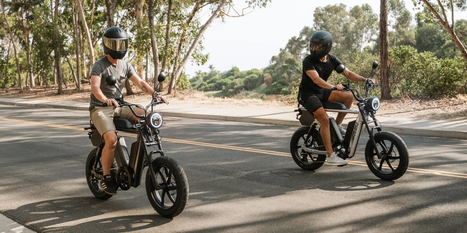 Two riders on electric bikes with helmets cycling through tree-lined road
