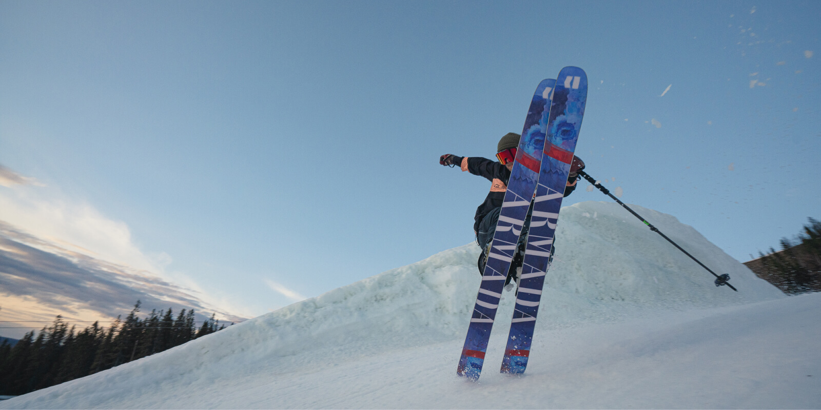 Skier mid-jump on snowy slope with blue skies and pine trees