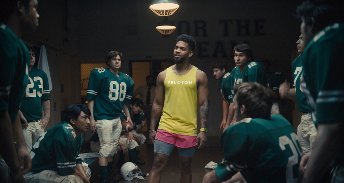 Peloton athlete in yellow tank top stands among football players in locker room