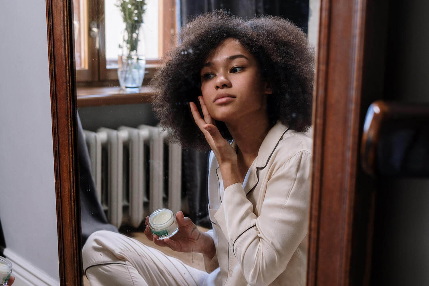 Person with curly hair examining face while holding skincare cream near window