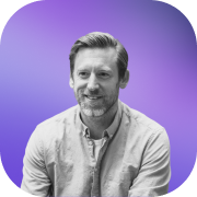Smiling man with short hair and beard wearing a light button-up shirt against a purple gradient background.