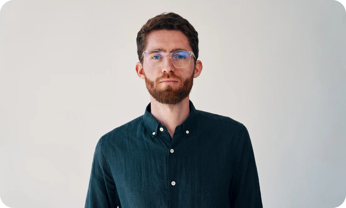 Portrait of Paul Becker, CEO and co-founder of re:cap, with reddish-brown hair, beard and clear glasses, wearing a dark green shirt against a light grey background.