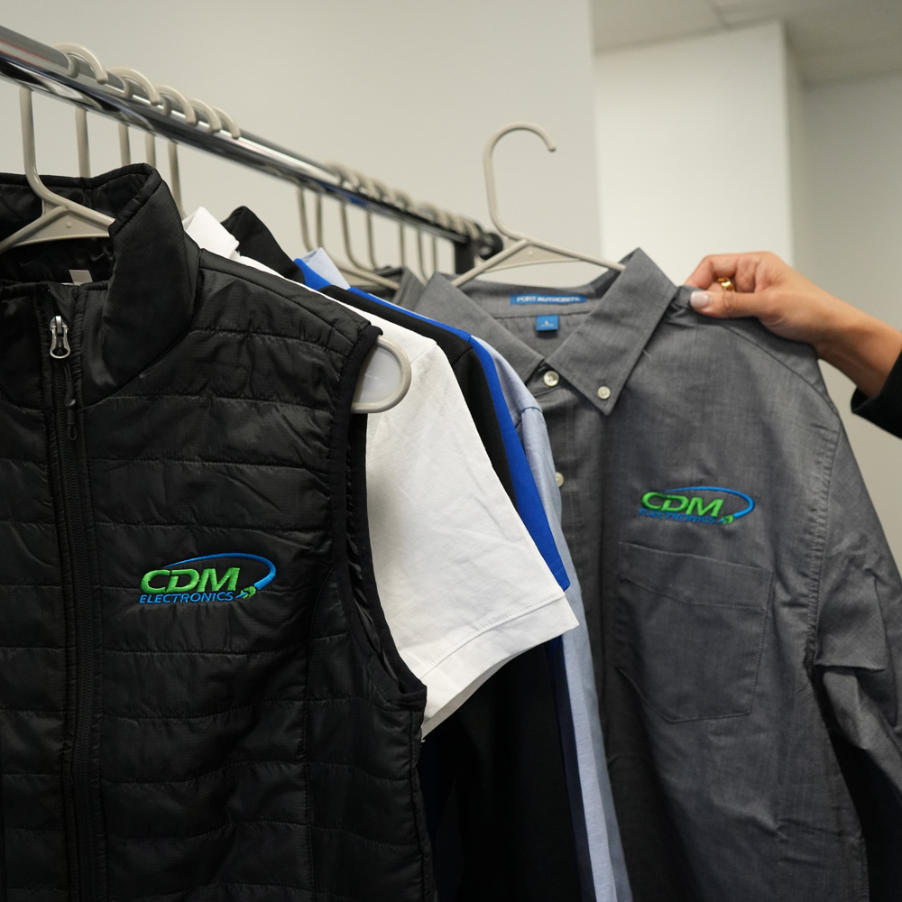 Black vest and gray button-up shirt with green and blue CDM Electronics logos hanging on rack among other shirts, with a hand holding the shirt collar.