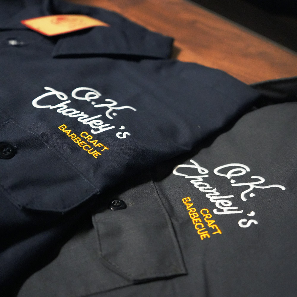 Two button-up shirts in dark colors with embroidered text reading C.K Charley's Craft Barbecue on the chest pockets.