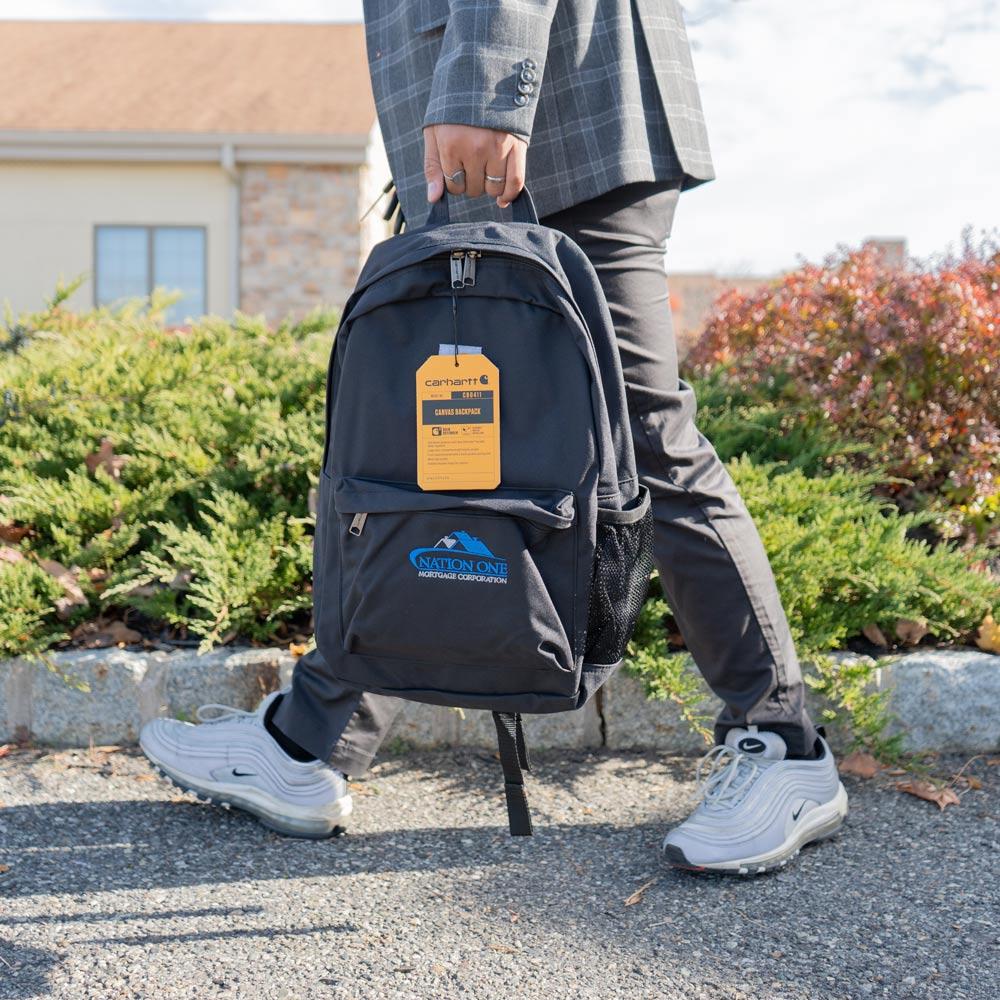 Person in gray plaid jacket and black pants walking on pavement holding a black Carhartt canvas backpack with Nation One Mortgage Corporation logo.