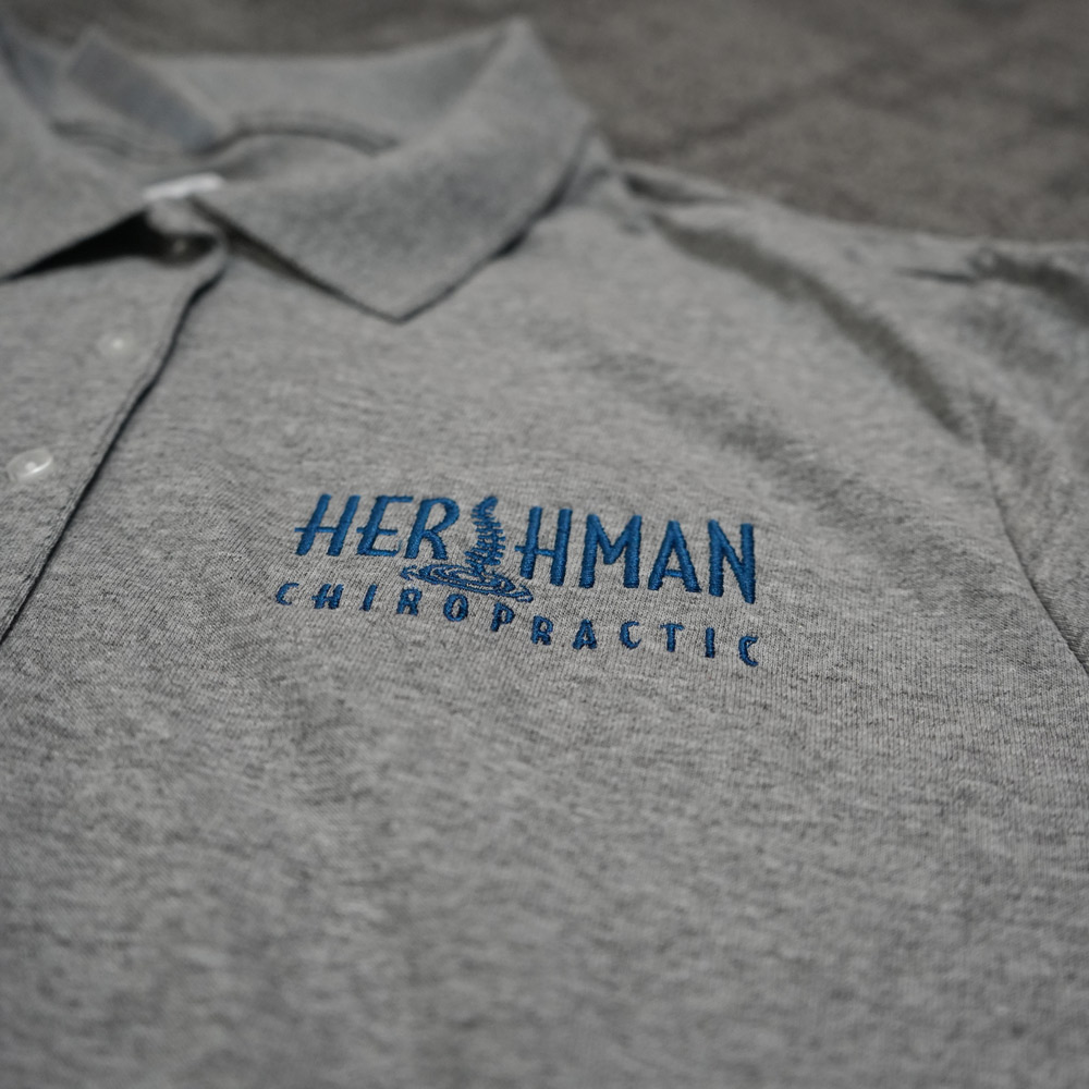 Gray polo shirt with blue embroidered text and spinal column graphic reading Hershman Chiropractic.