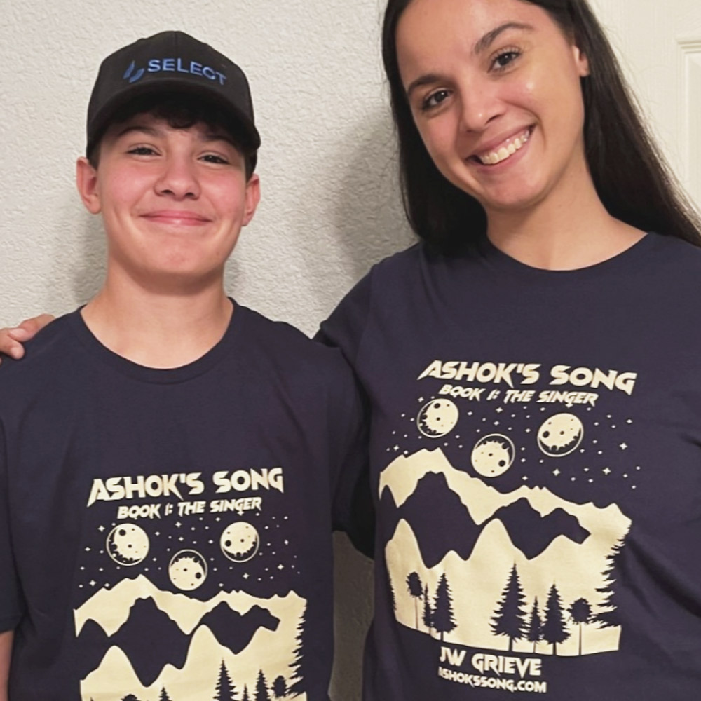 Two smiling people wearing navy blue t-shirts with a graphic of moon phases over mountains and trees, titled 'Ashok's Song Book 1: The Singer' by JW Grieve.