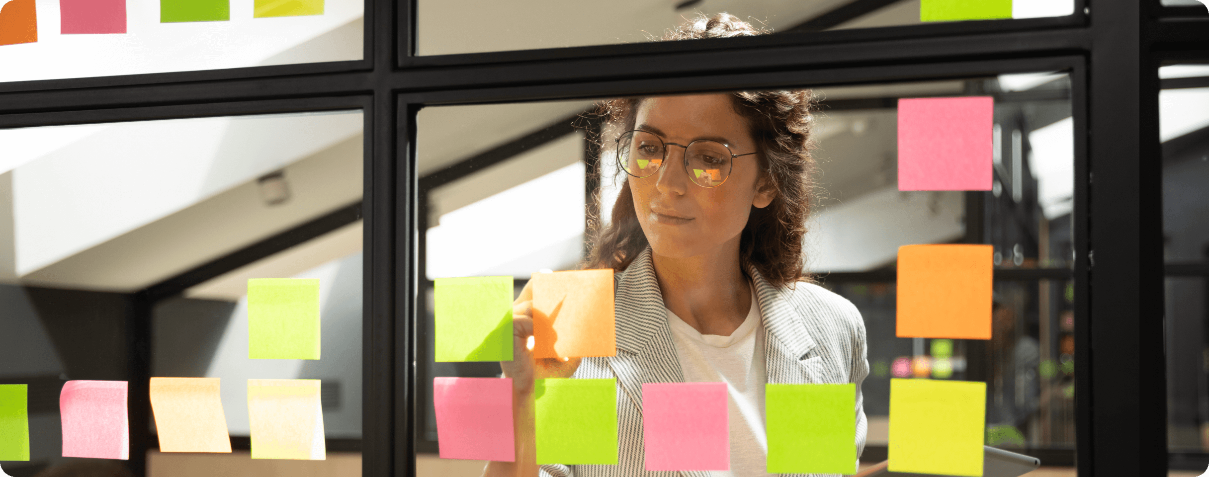 A woman wearing glasses stands in an office facing a wall of windows. She is rearranging bright green, yellow, and pink sticky notes that are stuck to the windows.