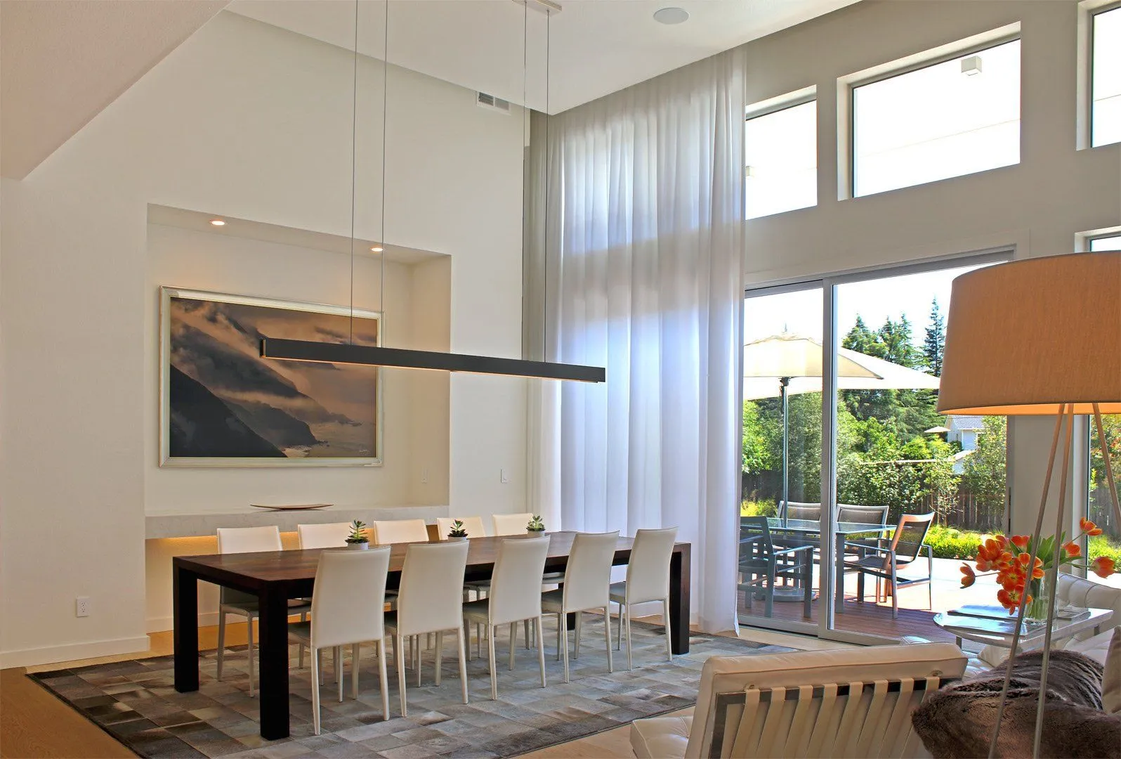 Dining room with sheer white drapes, tall windows, long table with white chairs, view of patio.