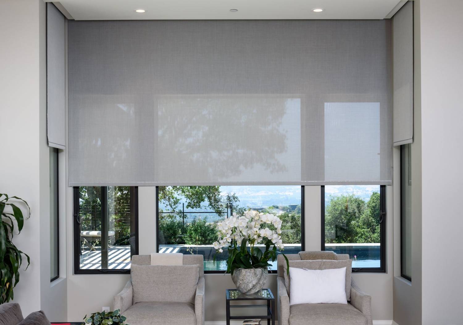 Modern living room with roller shades, large windows, and two armchairs facing a garden view.