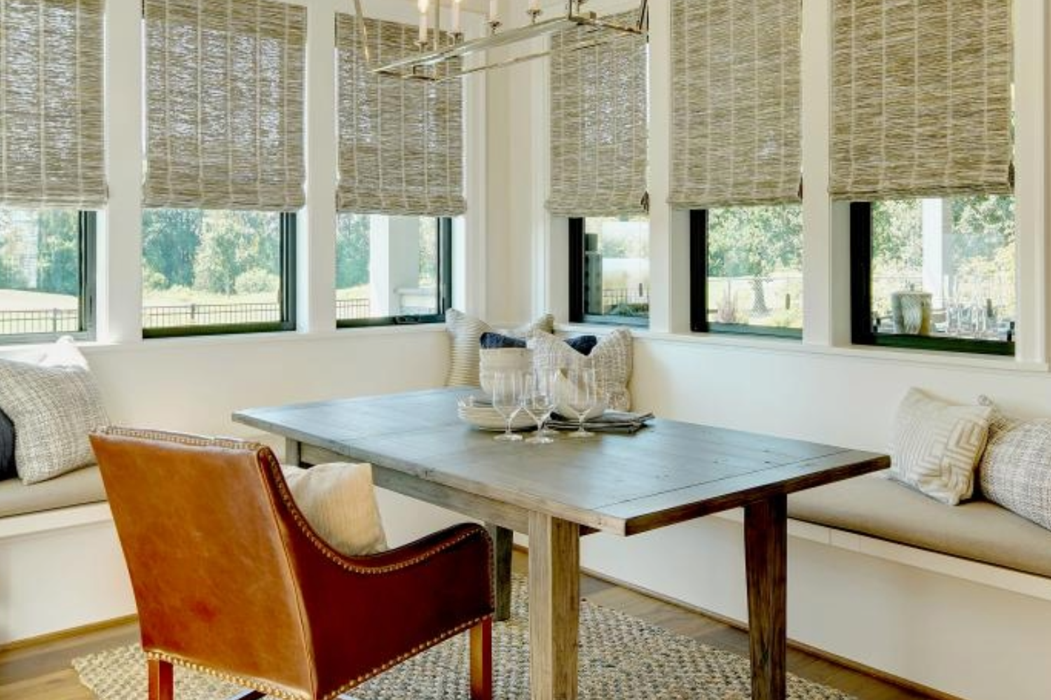 Sunlit dining nook with rustic wooden table, leather chair, and woven blinds overlooking a green landscape.