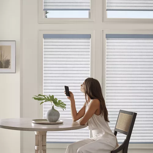 A woman in a white dress sits at a round table holding a smartphone, with large windows in the background and a plant vase on the table.