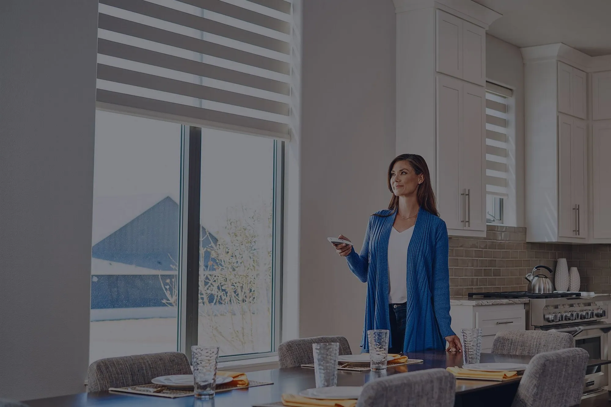 Woman in a blue cardigan using a remote to control motorized shades in a modern kitchen.