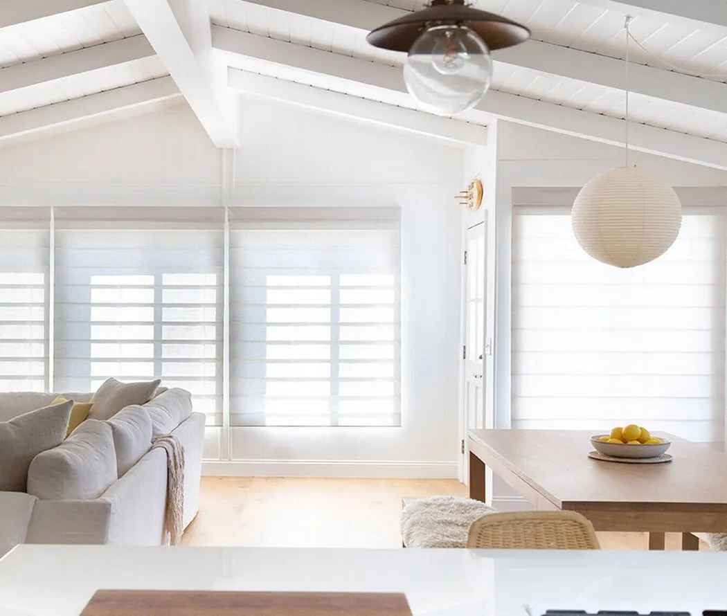 Bright open-plan living and dining room with motorized shades and exposed wooden beams.