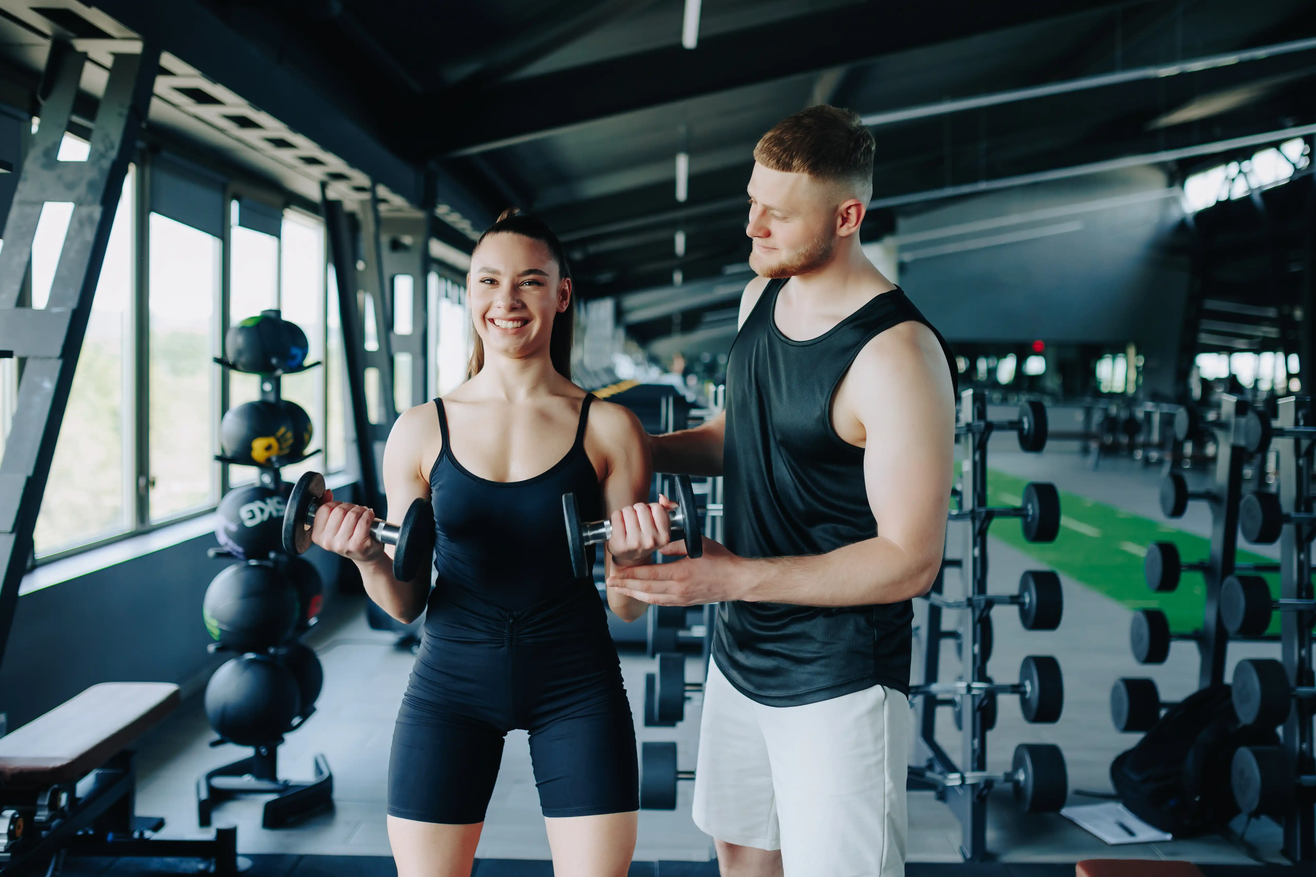 Smiling woman performing bicep curls with dumbbells while a personal trainer stands beside her to support her form.