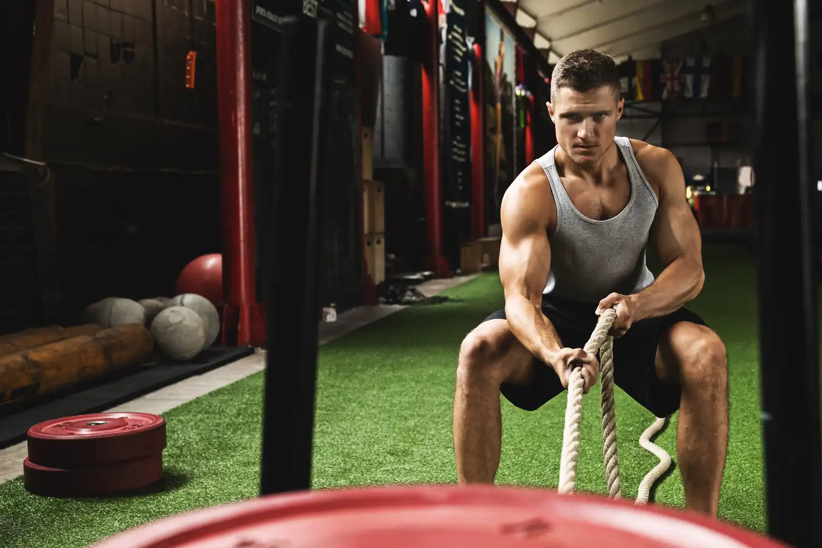 A muscular man wearing a grey tank top and black shorts crouching in a low squat on green artificial turf, gripping a thick white rope with both hands as he pulls a weighted sled toward him in a gym.