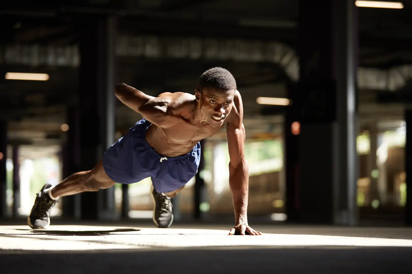 An athletic shirtless man performing a one-arm pushup in an outdoor or open gym setting