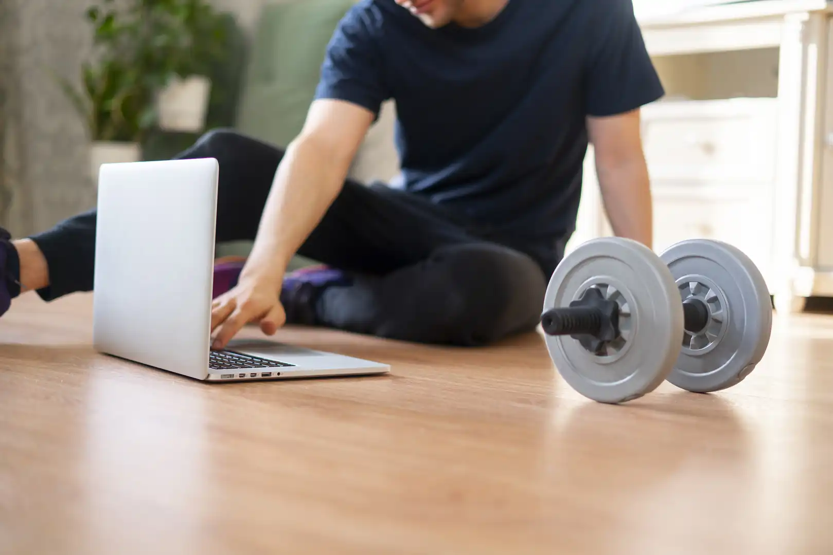 A man sitting on the living room floor typing on a laptop next to a adjustable dumbbell