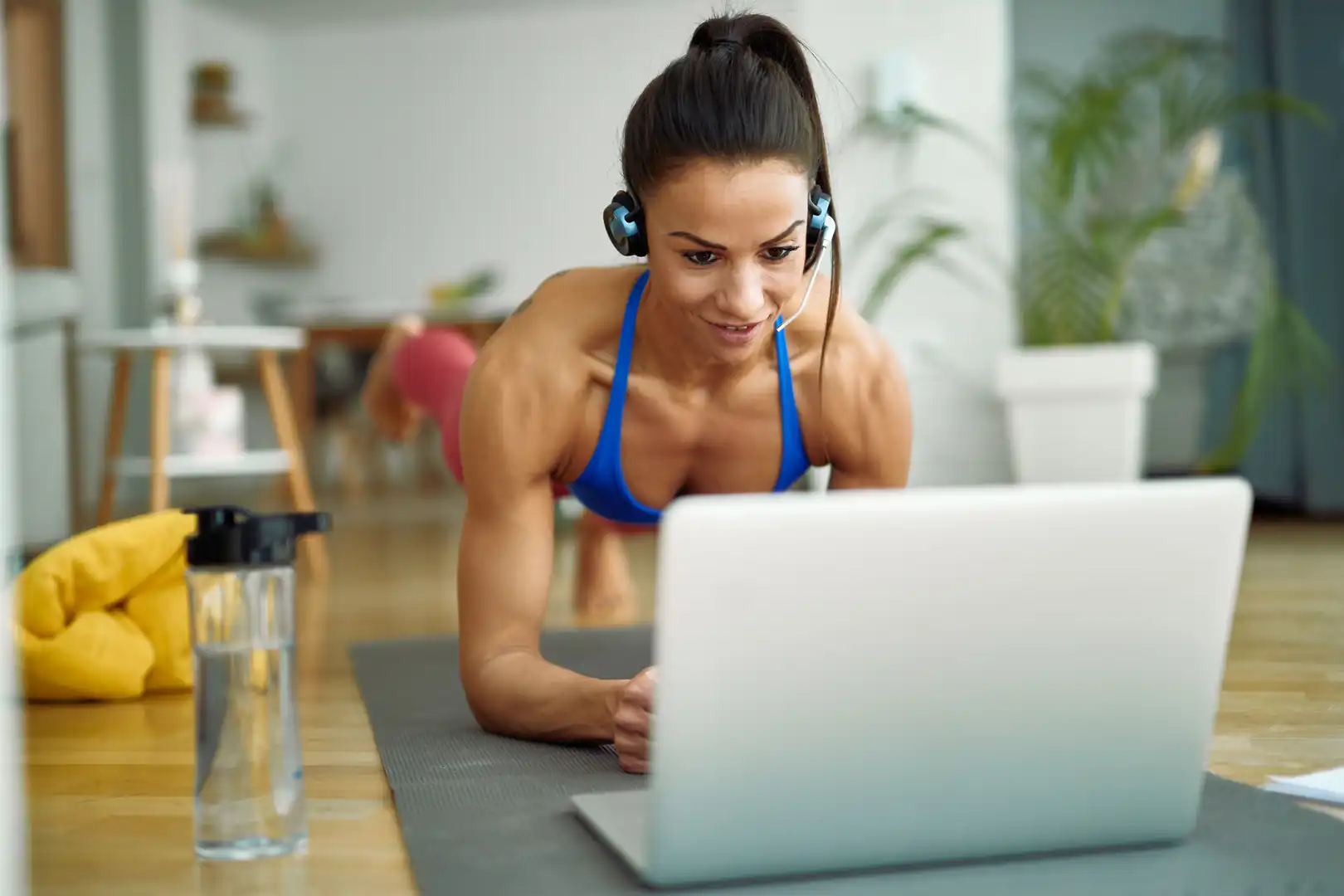 A muscular woman with her hair in a ponytail wearing a blue sports bra, pink leggings, and a headset with a microphone holds a forearm plank on a grey yoga mat while looking at a silver laptop in her living room.