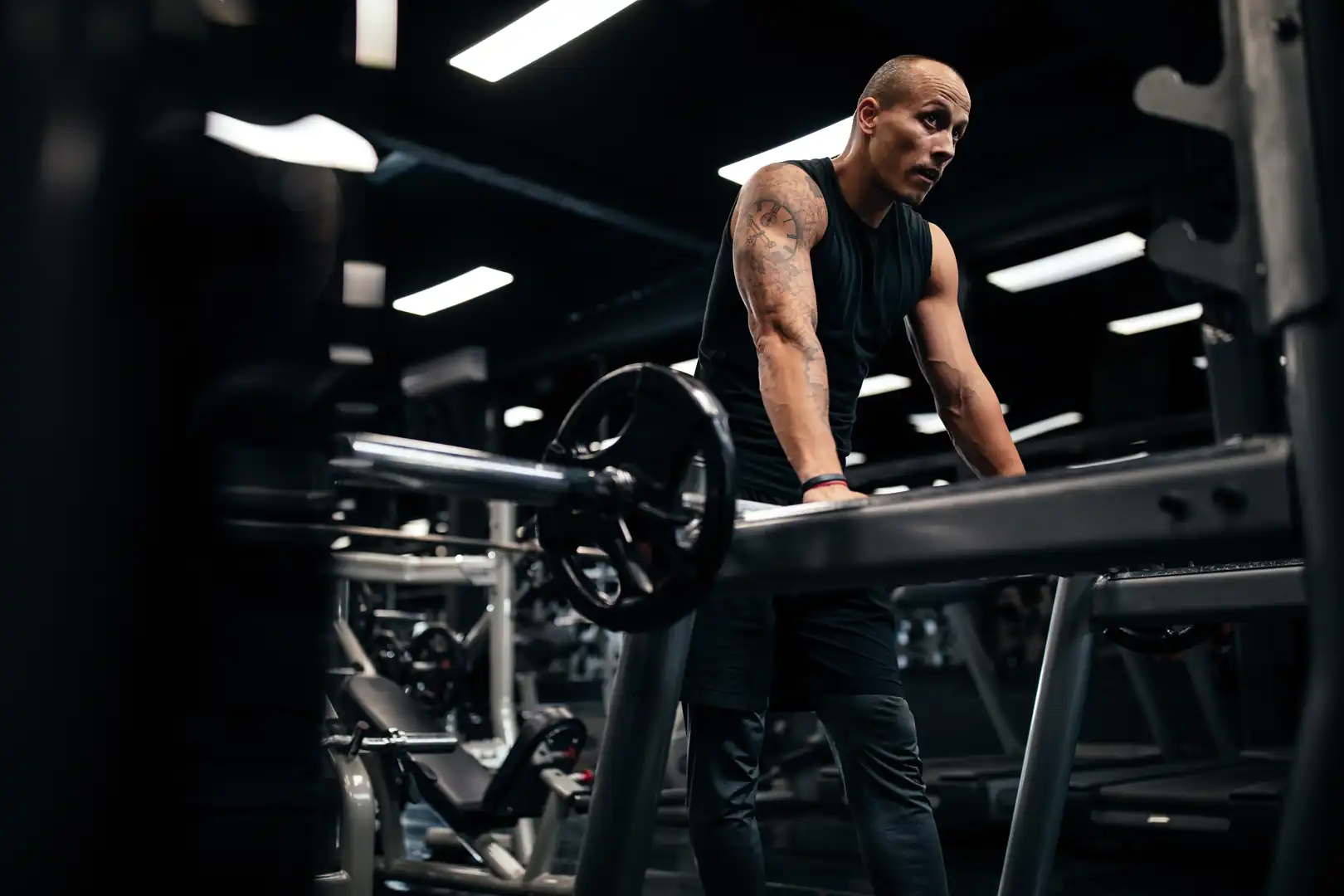 A muscular man with arm tattoos leaning against a barbell on a squat rack while taking a breath