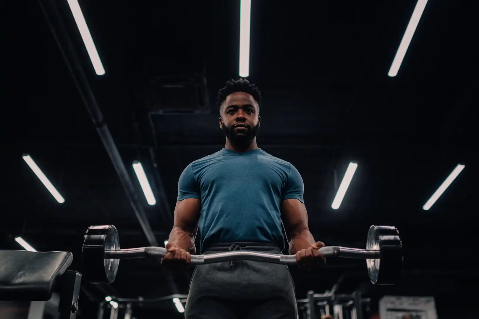 Low-angle shot of a determined Black man wearing a blue t-shirt and grey sweatpants performing a bicep curl with a weighted EZ curl bar in a dimly lit gym with overhead linear strip lighting.