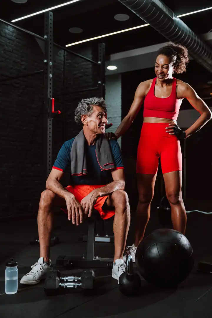 A senior man resting on a gym bench while smiling and conversing with his female personal trainer